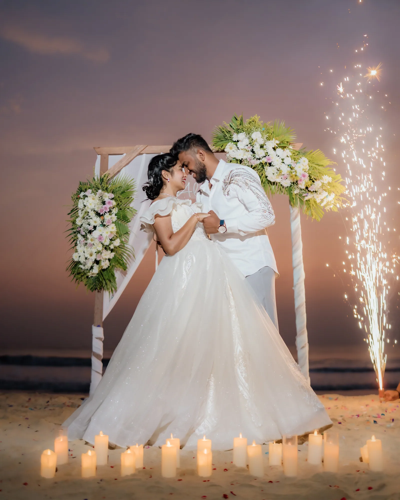 A couple under a flower arch by the sea, sparklers behind them — Darshan Ravi Photography